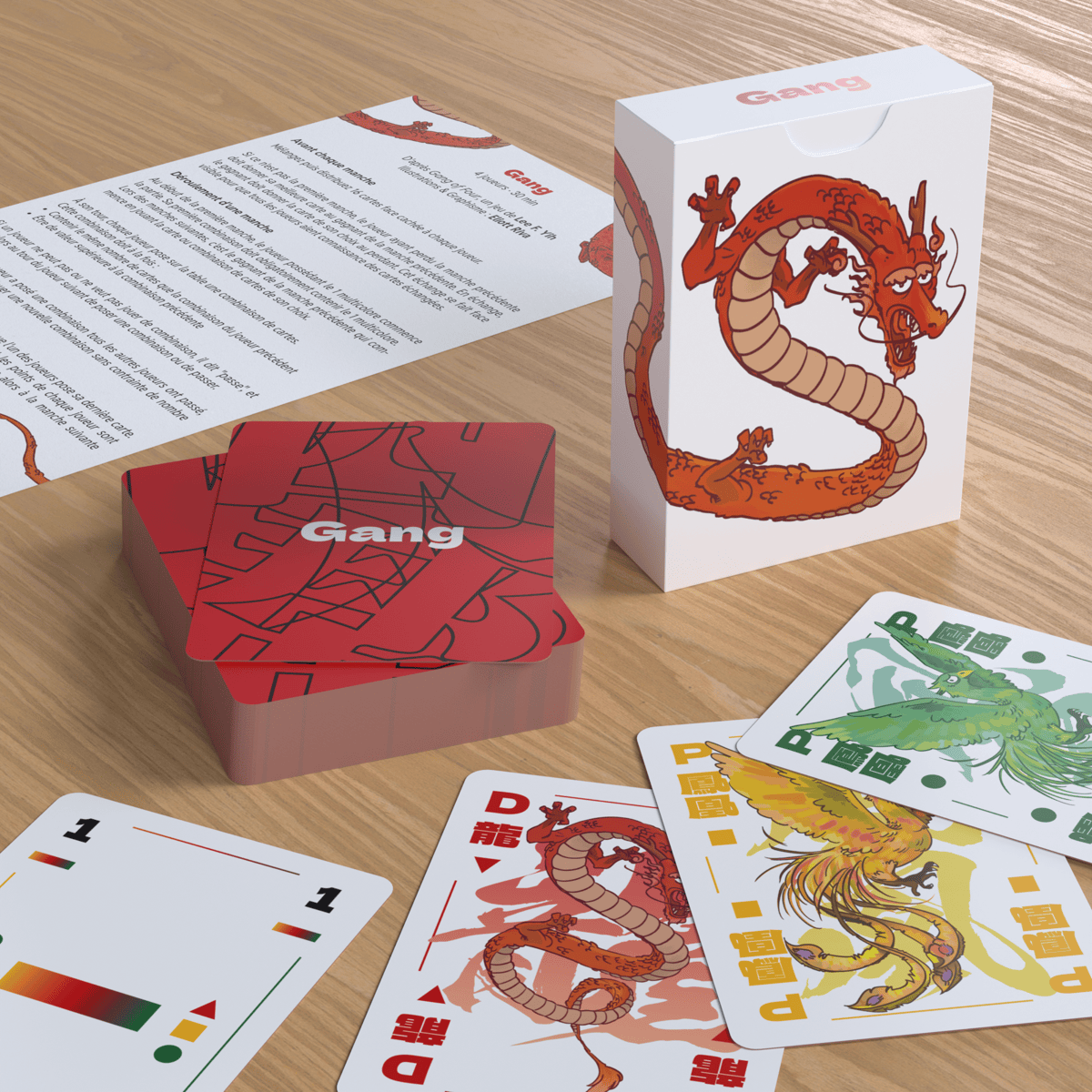 Realistic 3D view (like a photo) of all the elements of the Gang game arranged on a table. In the foreground, 4 cards are arranged in a disorganized fashion, as if they had simply been thrown onto the table. They are, from left to right, the multicolored 1, the dragon, the yellow phoenix and the green phoenix. A little further on, a pile of face-down cards can be seen, with the highest card at an angle, as well as the game's box, depicting a manga-style Chinese dragon, whose long red body runs around all sides of the box. Finally, in the background, a sheet of paper with the rules of the game printed on it sits on the table.